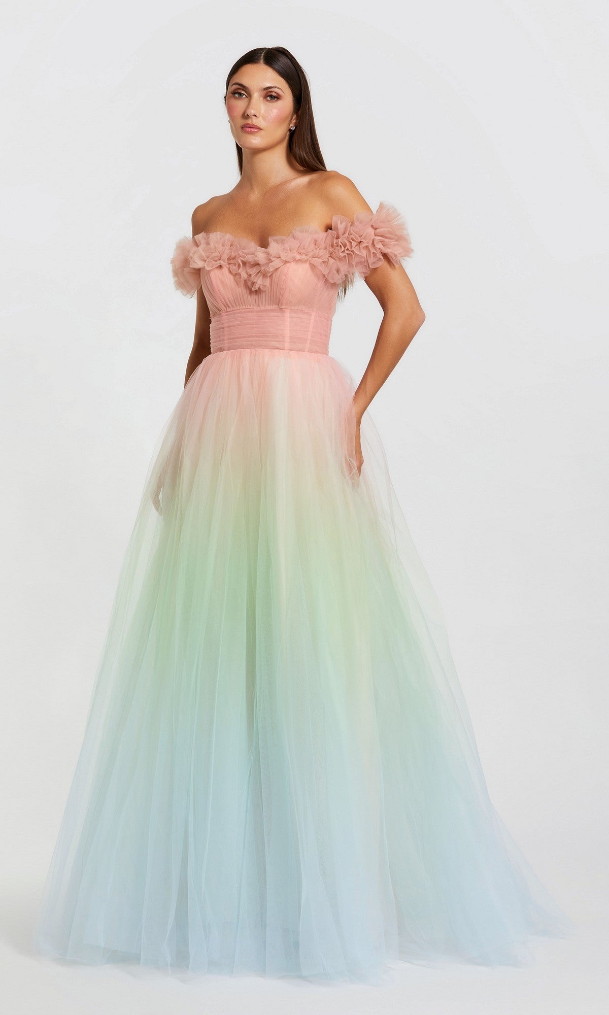 A woman in the Mac Duggal 37002 long prom dress in Pink Sorbet, featuring an off-the-shoulder design and pastel ombre tulle, stands against a plain light background.